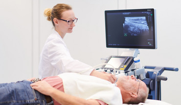 Eine junge Ärztin führt bei einem älteren Patienten eine Ultraschalluntersuchung der Schilddrüse durch.